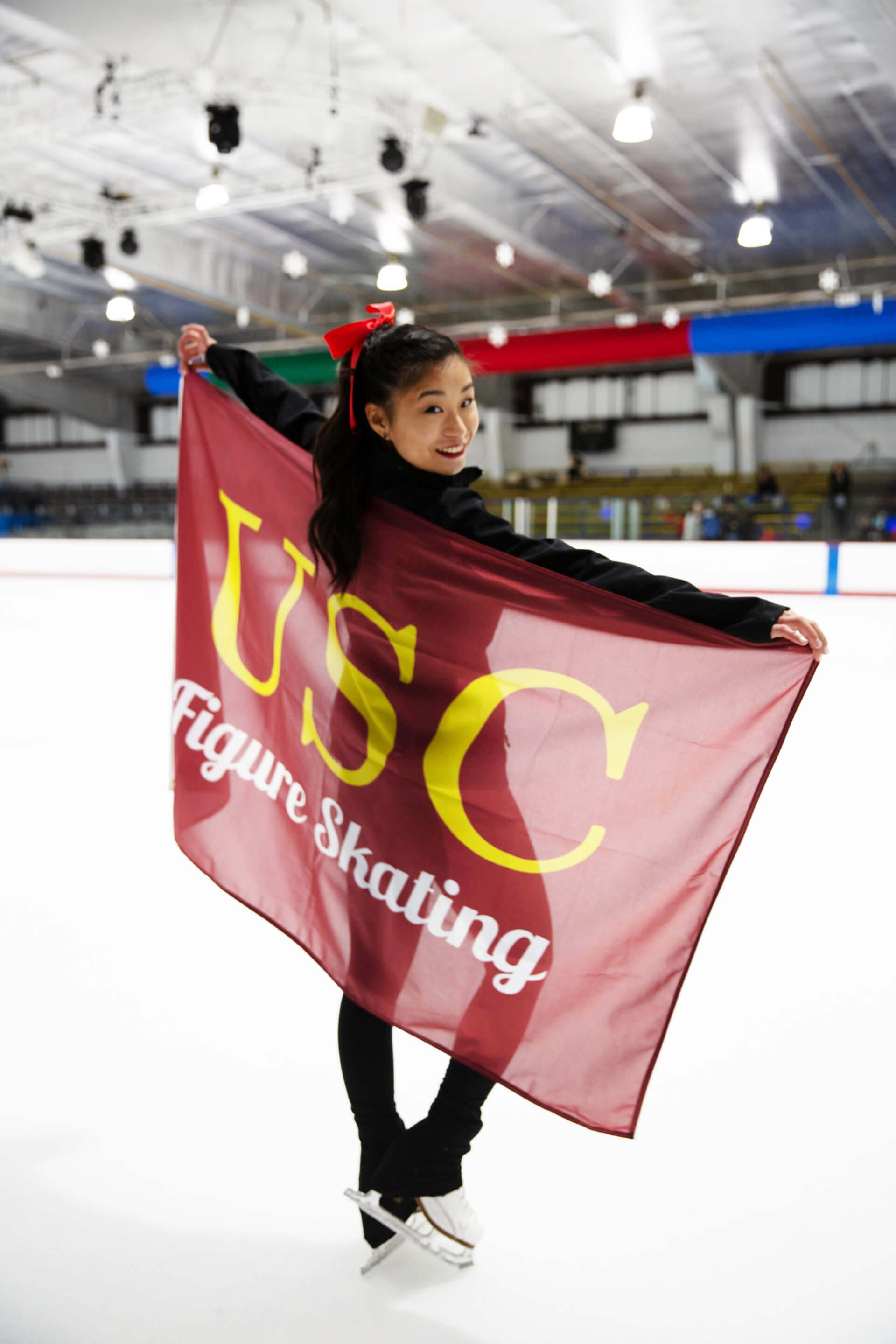 Nicole Wong, who graduated from USC Viterbi in 2019, pictured on the ice with her figure skates. Looking over her right shoulder, she holds a flag that says USC Figure Skating, which hangs over her back. 