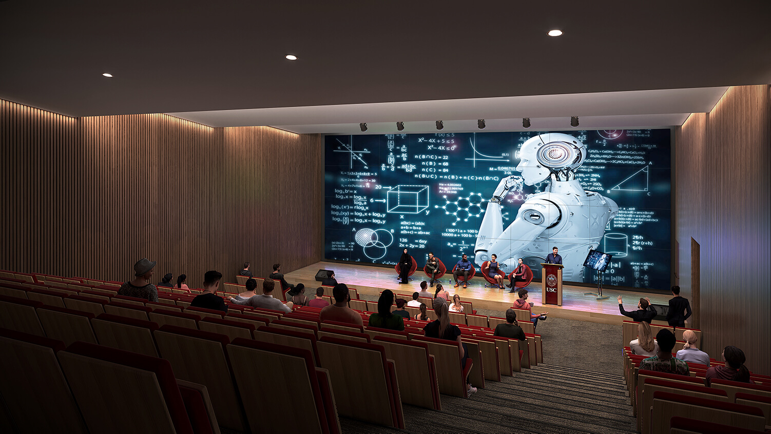 A large auditorium, with people sitting on the stage and an image of a robot projected along the wall behind the stage.