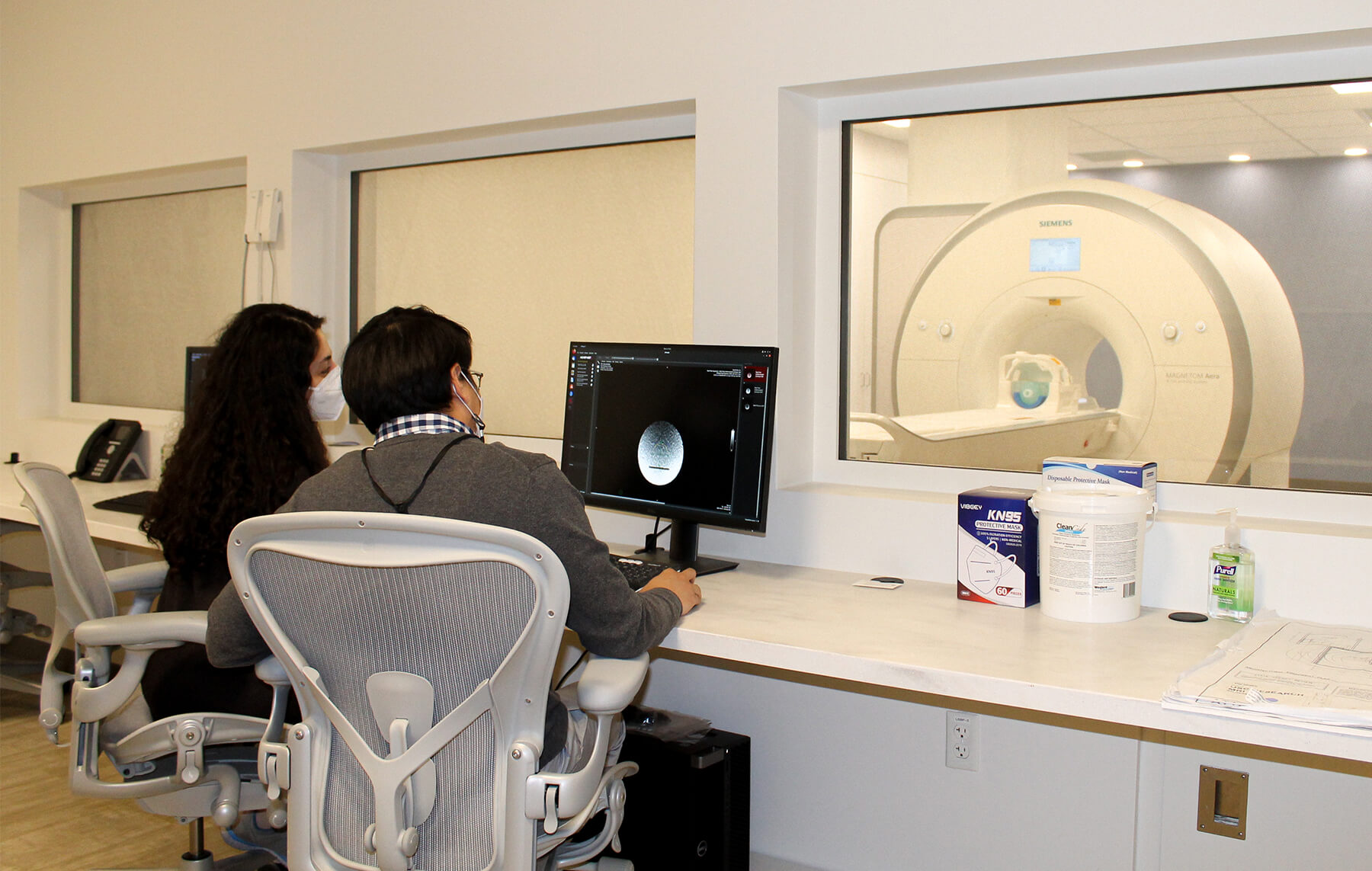 Two researchers sit facing away from the reader from the monitoring room next to the MRI. They are in front of computers looking at the images the MRI machine in producing.  